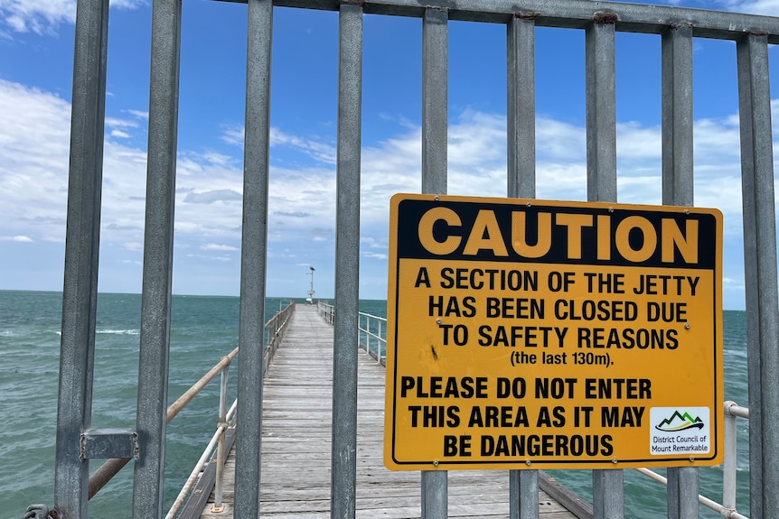 A silver barred gate shuts off a part of a wooden jetty, with a yellow sign reading 'CAUTION' 