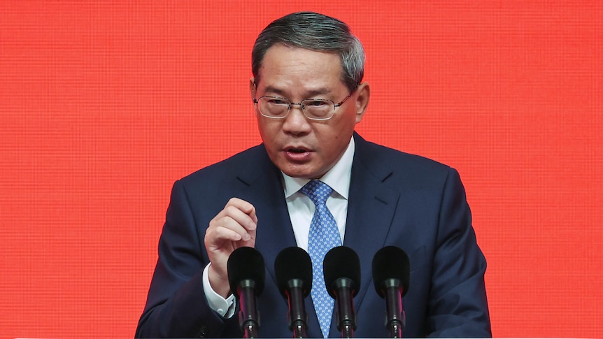 A man in a suit and tie stands behind a bank of microphones speaking at a press conference