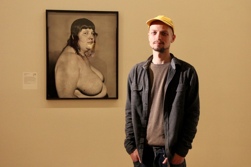 A man in a yellow cap stands in front of a wall with a black and white image of a topless person hung on it.