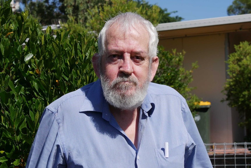 An elderly man with a grey beard, wearing a blue shirt standing outside looking into camera.