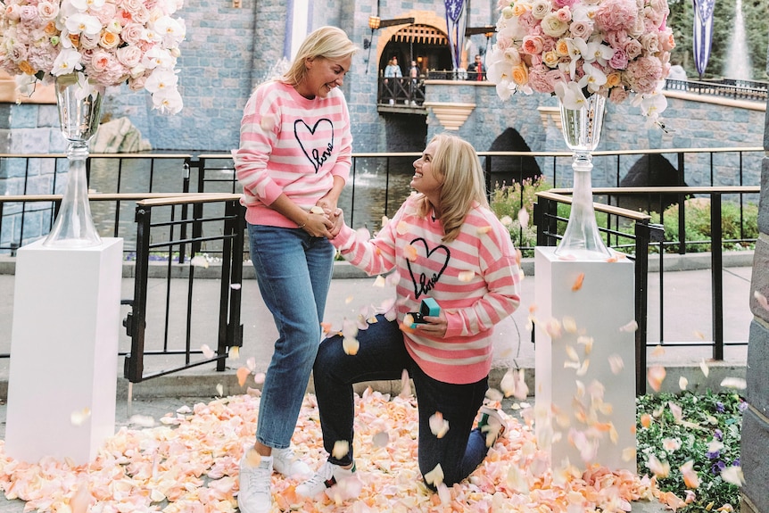 Rebel Wilson wearing pink and white on one knee proposing to Ramona Agruma in matching clothes at Disneyland