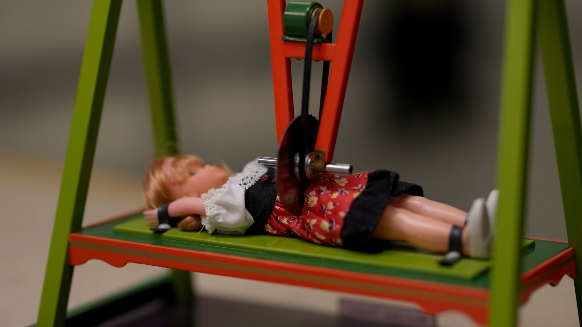 A small doll lies on a miniature table, with a tiny saw cutting her in half through her stomach