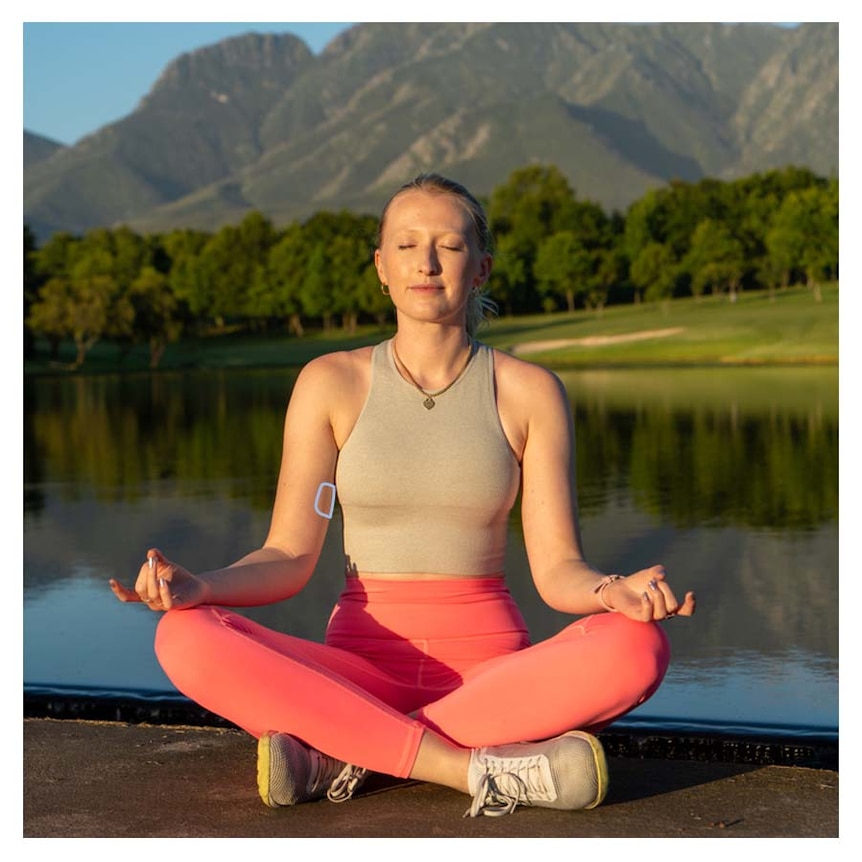 A young woman wearing pink exercise wear meditating with her eyes closed, you can see a patch on the inside of her right arm