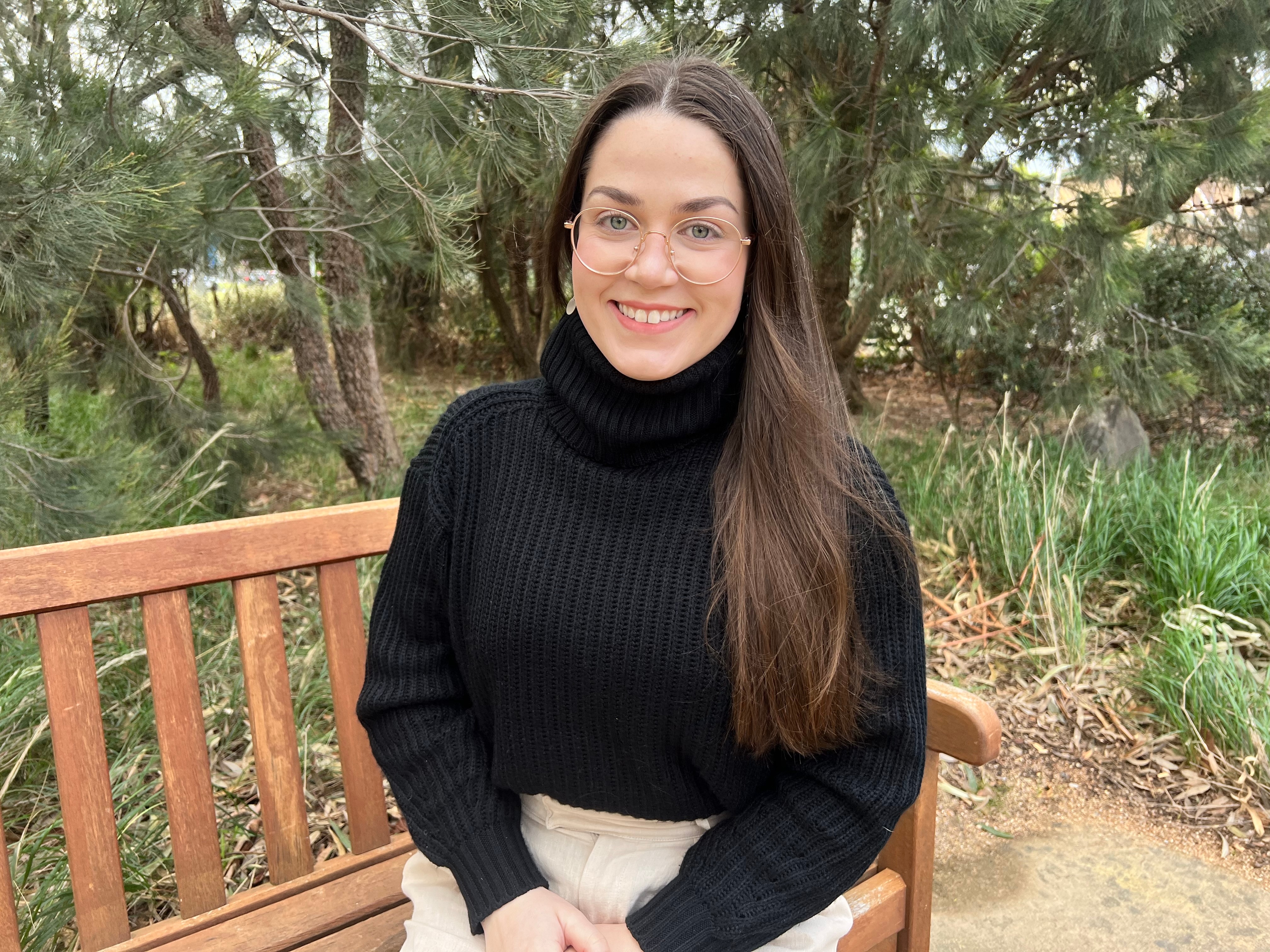 Young woman in black high neck jumper, wears glasses, smiling, long brown hair, sitting on park bench with trees behind her