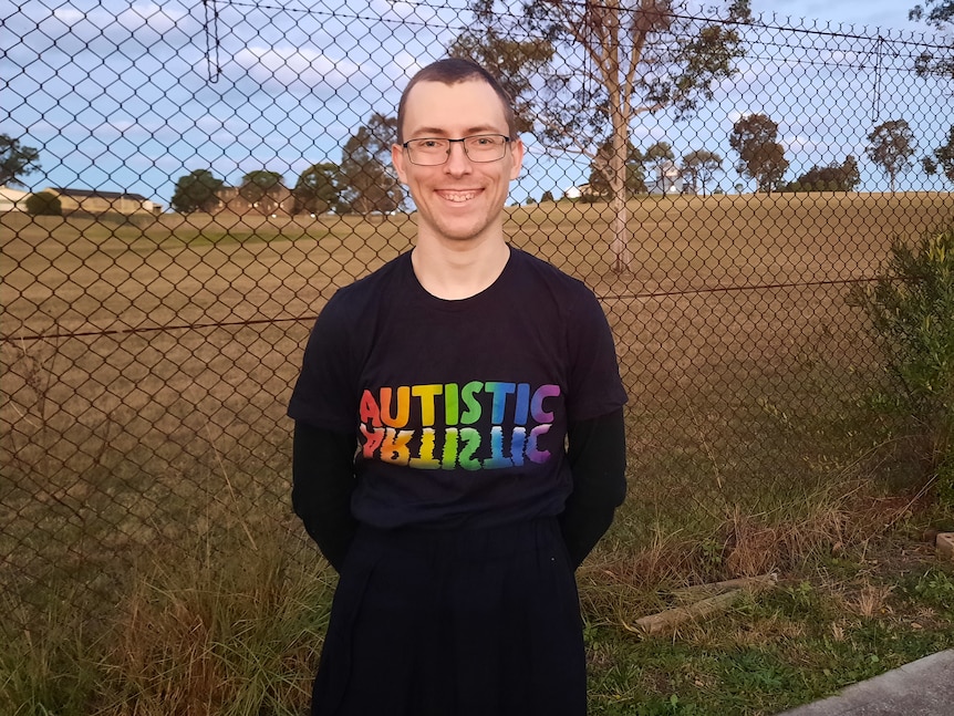 Brendan smiles standing in front of a field, wearing a shirt that says 'autistic' in rainbow letters.