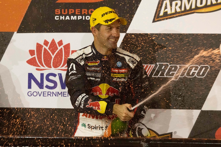 Le pilote Jamie Whincup sourit en giclant du champagne sur le podium des gagnants avec des pancartes publicitaires derrière