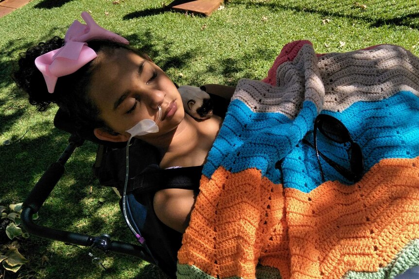 Denishar Woods, wearing a pink bow in her hair, lies in a wheelchair on grass with a tube attached to her nose.