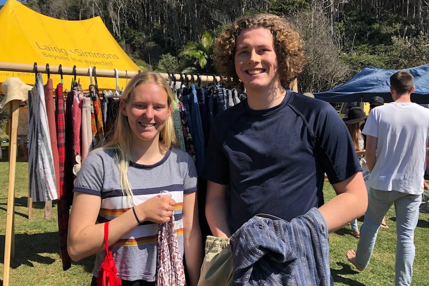 Two people standing holding clothes in front of a clothing rack at markets.