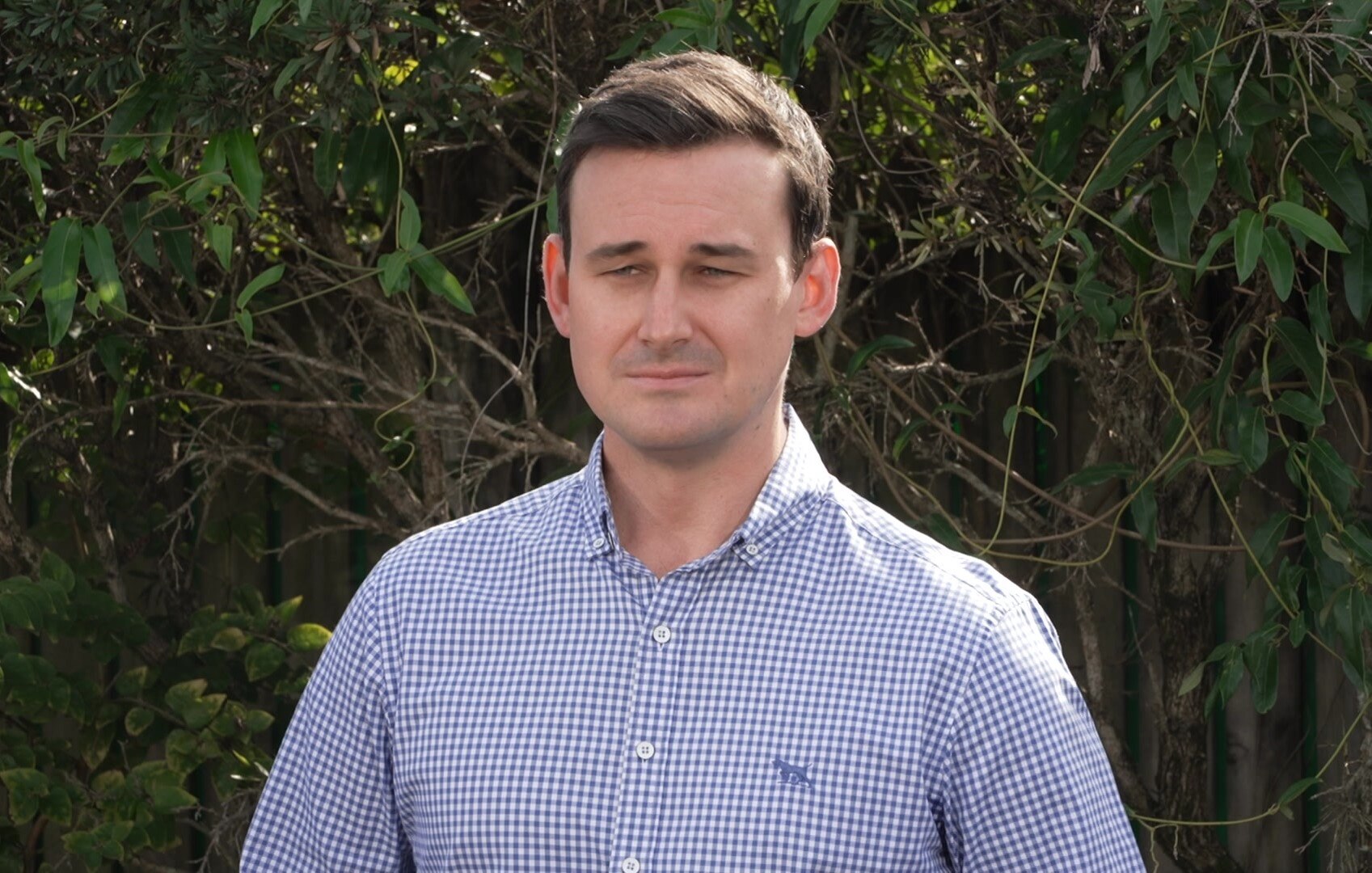 A man in a blue and white checkered shirt standing in front of greenery
