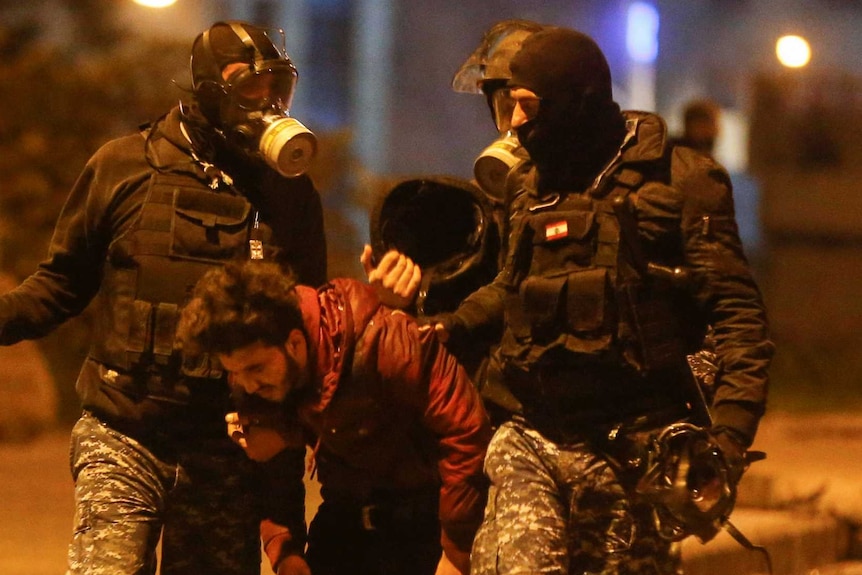 Three police in riot gear wearing gas masks hold a man as he bends over.