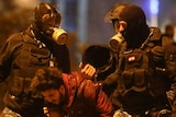 Three police in riot gear wearing gas masks hold a man as he bends over.
