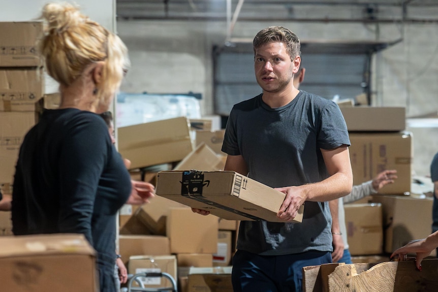 A young man in grey shirt hands a cardboard box to a blonde woman with back to camera