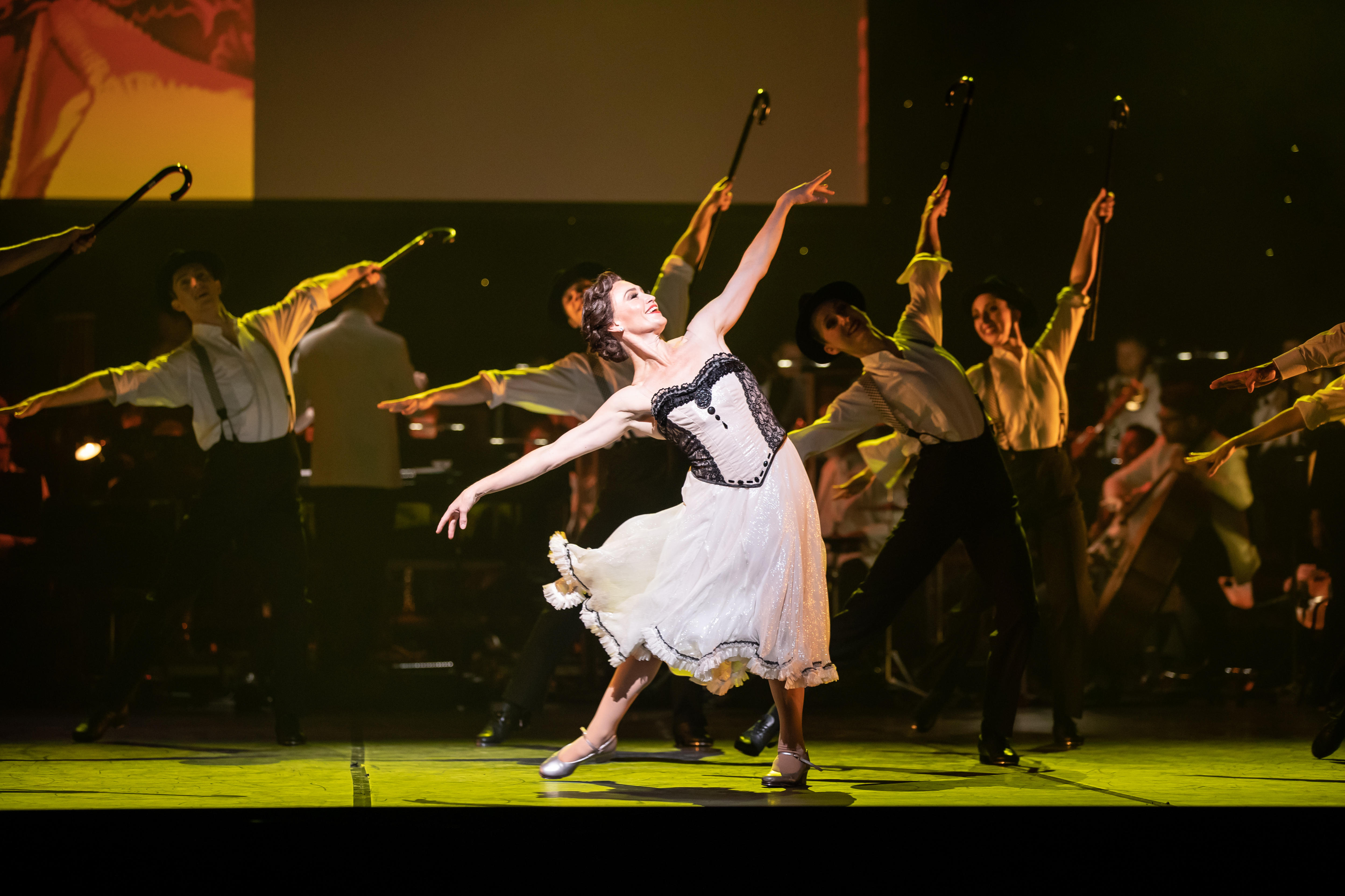 Woman in white dress dances on stage with back up men dancing behind her