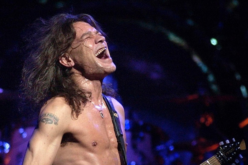 A shirtless long-haired man plays guitar while singing.