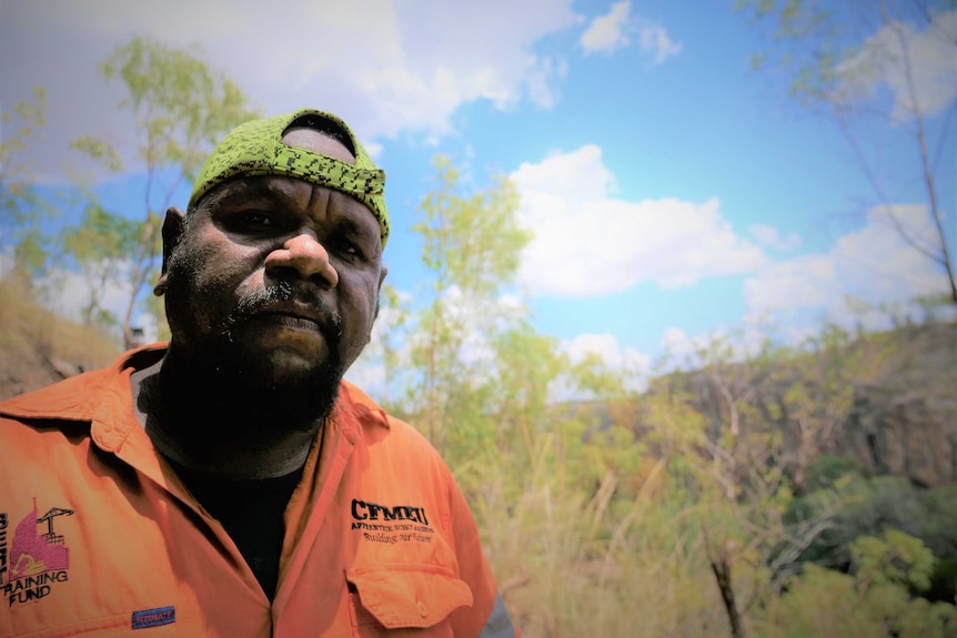 Man looking at camera wearing hi-viz and a green baseball cap backwards. Blue sky behind and bush setting