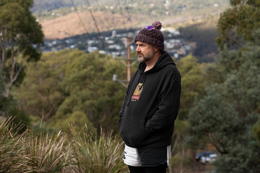A despondent looking man with a beard stands on a hillside wearing a beanie. 