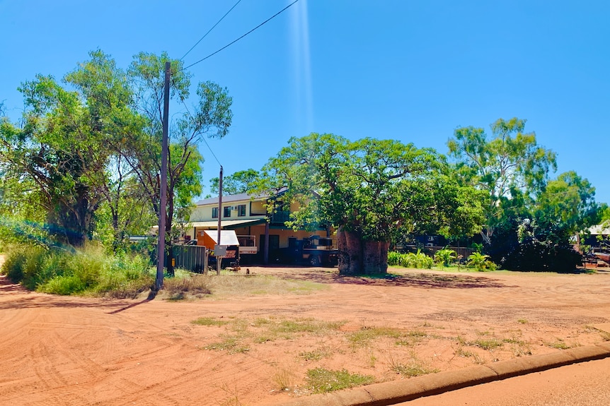 A house behind trees with a wide expanse of red dirt in front.