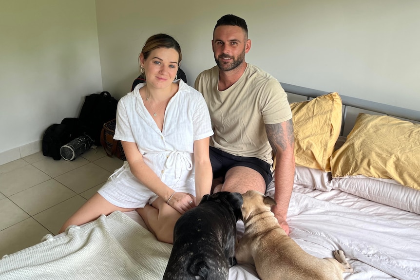 A young couple with two dogs sit on a bed. They are smiling at the camera.