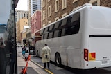 A bus in a laneway with old stone buildings and people wearing PPE