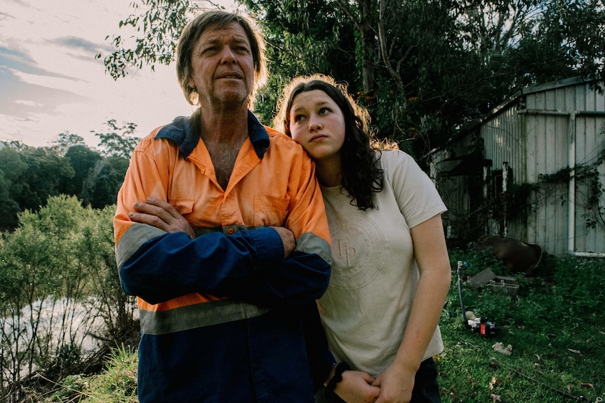Mark O'Toole stands with arms folded in a garden as his daughter Eliza rests her head on his shoulder.