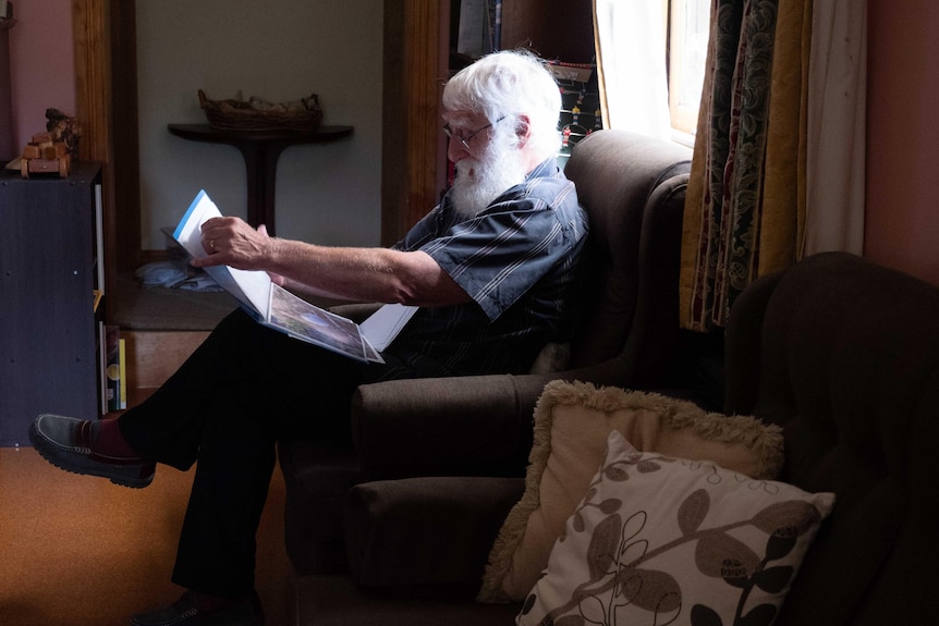 A man with a white beard sitting on a couch looking at a folder of photos