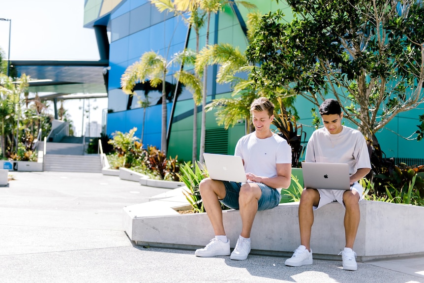 Two male students using laptops on bench at university.