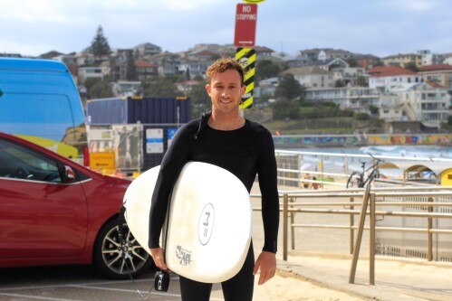 A man in a wetsuit standing with a surfboard in front of a beach.