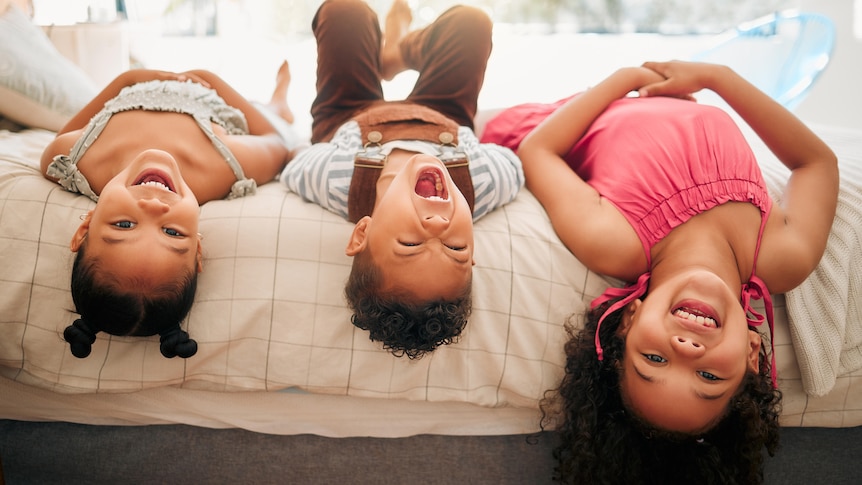 Three siblings lie upside down on a bed, laughing and looking at the camera.