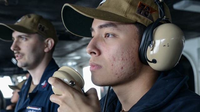 A sailor with earmuffs and a cap speaks into the radio onboard the ship.