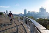 Cyclist wearing mask rides near fence of lookout over looking Perth city skyline of tall buildings. 