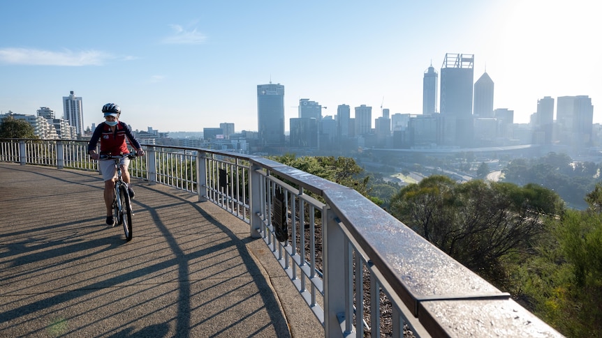 Cyclist wearing mask rides near fence of lookout over looking Perth city skyline of tall buildings. 