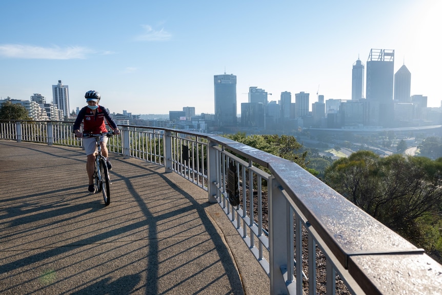 Cyclist wearing mask rides near fence of lookout over looking Perth city skyline of tall buildings. 