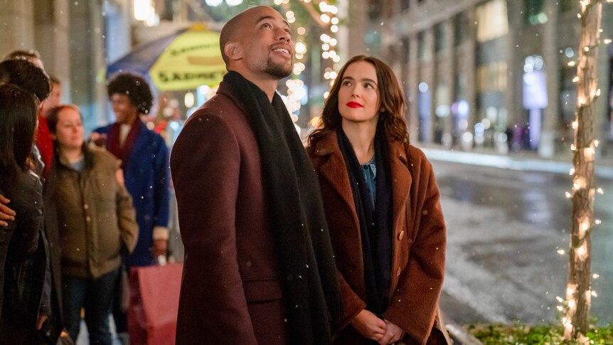 Kendrick Sampson, left, stands next to Zoe Deutch and looks up as she looks at him while they're in a fairy light lit street.