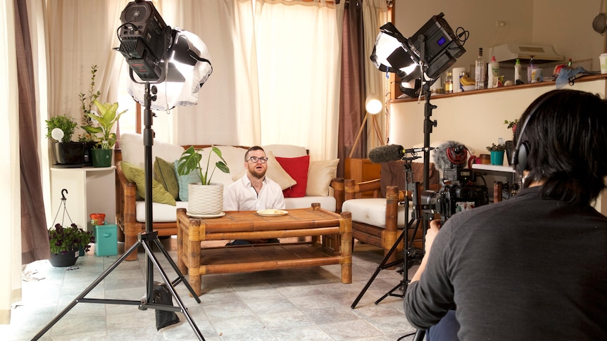 Angus is sitting on the floor behind a coffee table. There are lights on stands above him and a film camera pointed at him