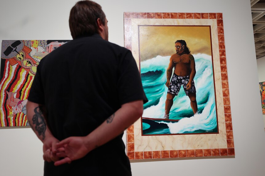A man in a black shirt stands in front of a painting of an Aboriginal man on a surfboard