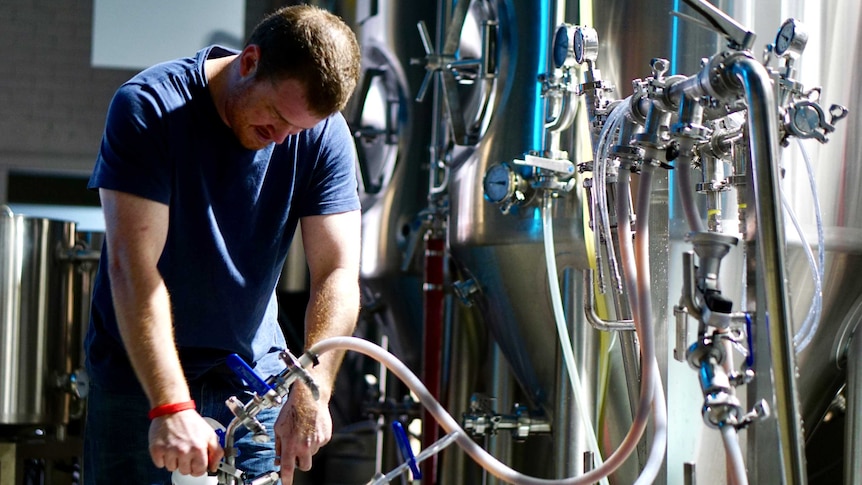A man in a t-shirt attaches pipe to a keg of beer, surrounded by brewing equipment.