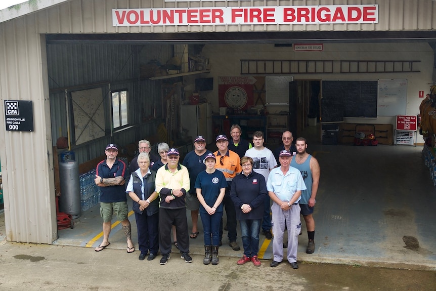 A group of people stand with hands clasped in front of them look up at the camera with a large garage behind them.