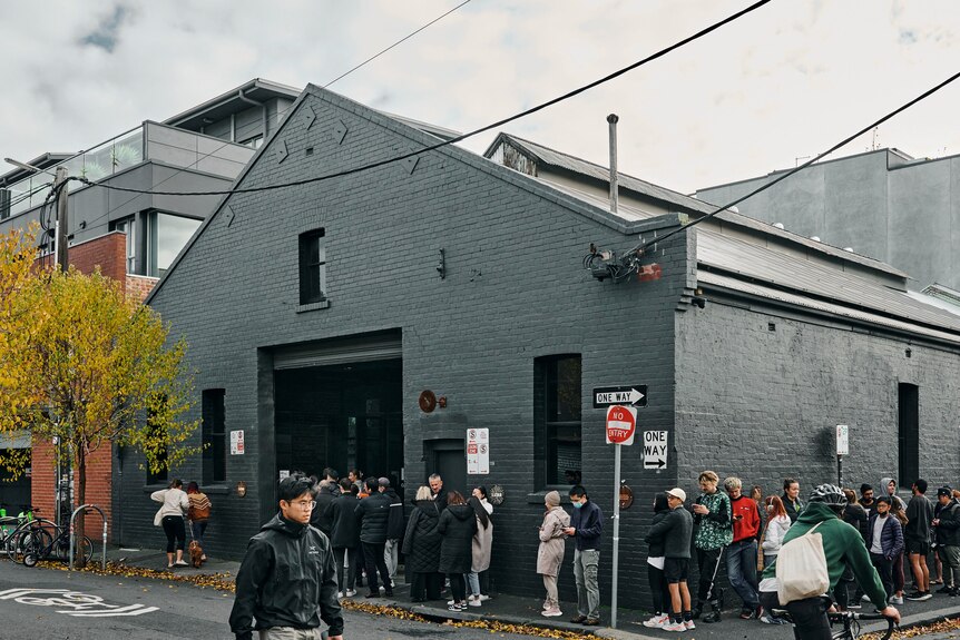 A line of people outside a grey warehouse
