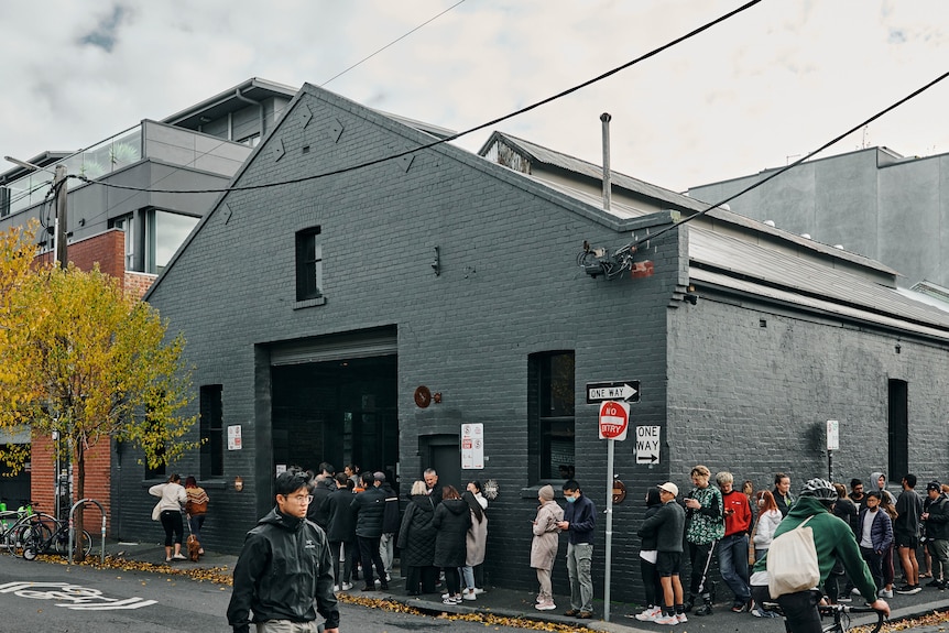 A line of people outside a grey warehouse