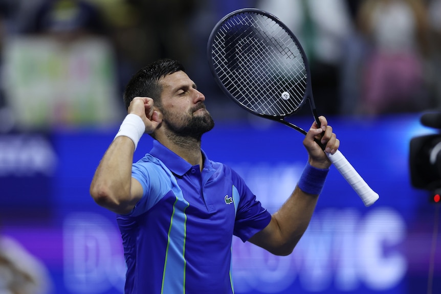 A male tennis player in a blue shirt, holdsn his arms up in victory, with a racquet in one hand.