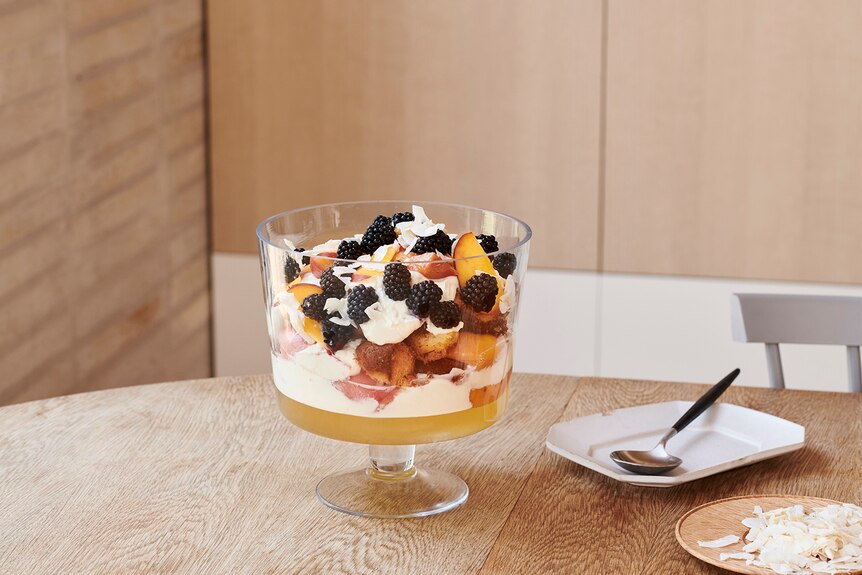 A large layered fruit trifle in a glass bowl on a timber dining table, toasted coconut and serve ware in the background.