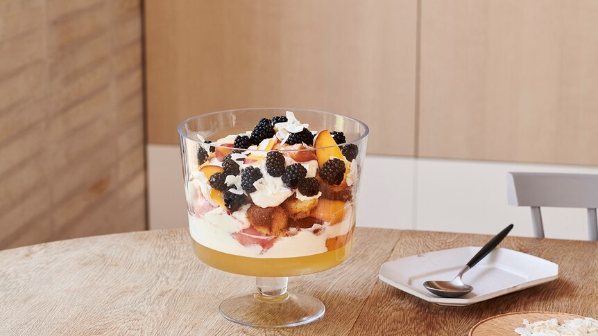 A large layered fruit trifle in a glass bowl on a timber dining table, toasted coconut and serve ware in the background.