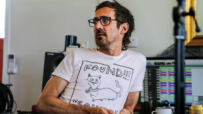 Gareth Liddiard wearing black-framed seeing glasses looks across room, sitting in front of production computer.