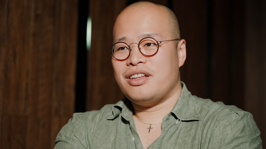 A close up photo of a smiling man wearing red glasses, a cross necklace and a green shirt.