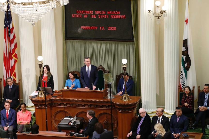 A man dressed in a suit gives a speech to a seated audience within a room.