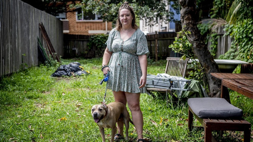 A young white woman with a green dress, standing in an overgrown backyard with a staffy dog