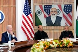Three men sit at a table in front of a large monitor displaying another man with Indian and US flags visible