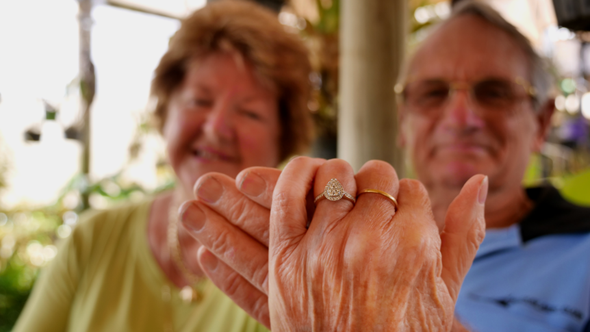 An older woman and an older man sit in a garden, holding up her hand with an engagement ring on it 