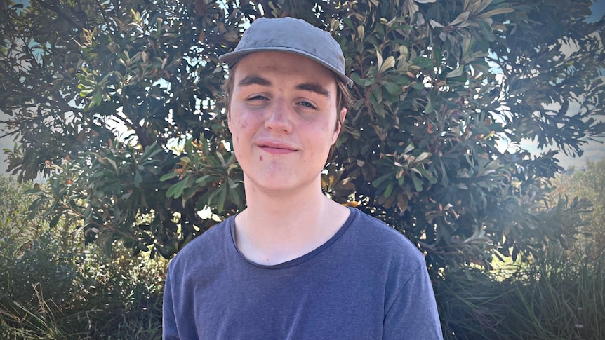 Teenage boy wearing a blue shirt and cap standing in front of a tree. 