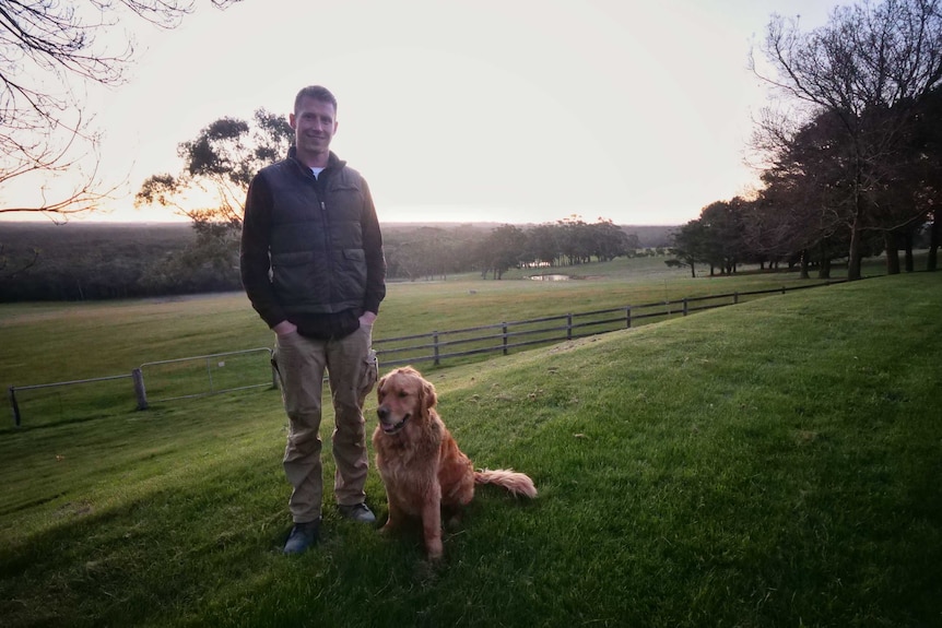 Dylan Grimes, wearing a jacket, smiles at the camera on a farm with his Golden Retriever dog.
